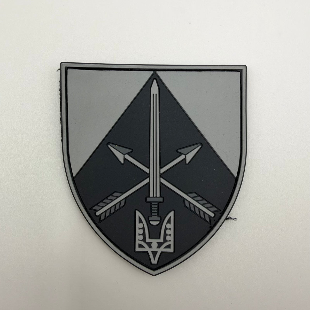 Grey and black shield-shaped patch featuring an upright sword over crossed arrows, with a stylised two-pronged emblem at the base.