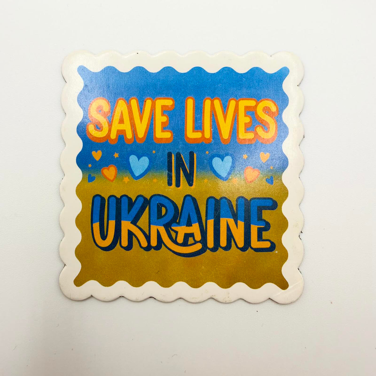 Fridge magnet reading “Save Lives in Ukraine” with colourful hearts and stars, a blue and yellow background and a cream scalloped border.