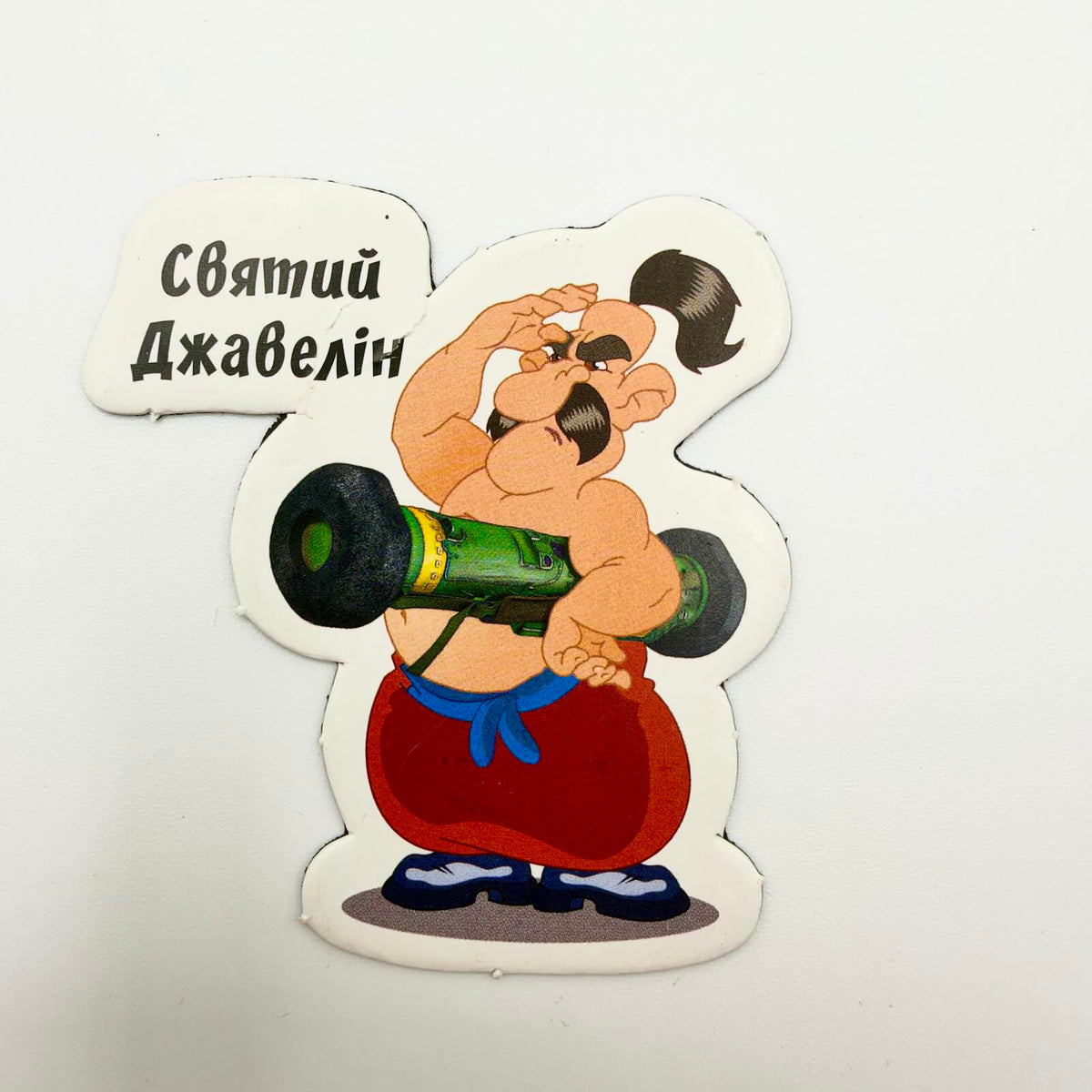 Shaped fridge magnet depicting a cartoon Cossack in red trousers carrying a green launcher, with Ukrainian text meaning “Saint Javelin”.