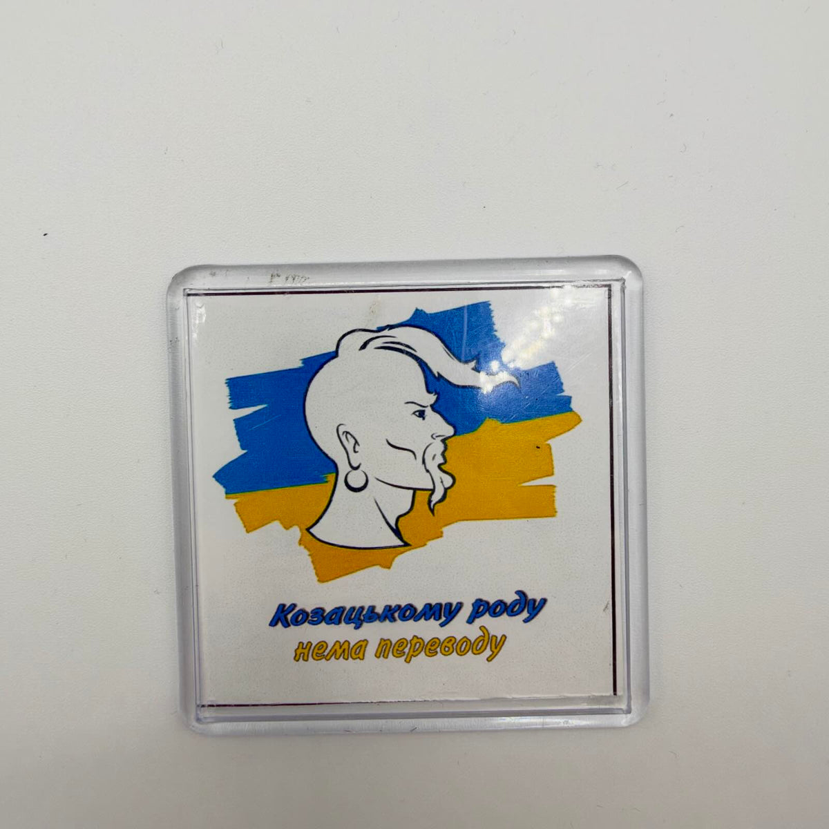 Ukrainian fridge magnet in a clear casing, showing a Cossack portrait over blue and yellow brushstrokes with the words “The Cossack line will never die out” in Ukrainian.