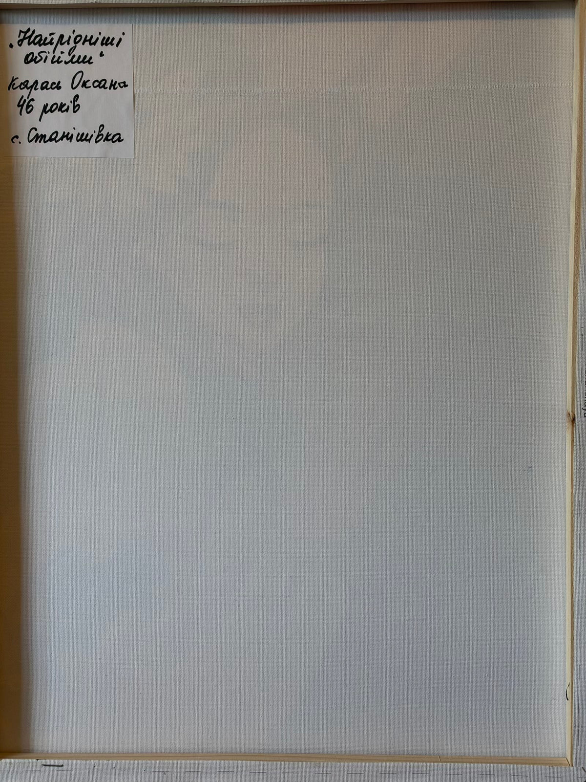 Reverse of the canvas showing its wooden stretcher frame and a handwritten Ukrainian label in the upper-left corner. The label gives the title “The Dearest Embrace” and identifies the artist as Oksana Karas, aged 46, from Stanishivka village.