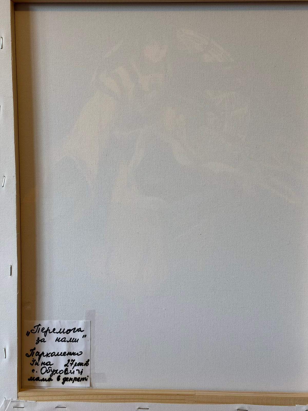 Reverse of the canvas showing its wooden stretcher frame and a handwritten Ukrainian label at the lower left. It reads “Victory Is Ours”, Inna Parkhomenko, aged 27, Obukhovychi village, a mum on maternity leave.