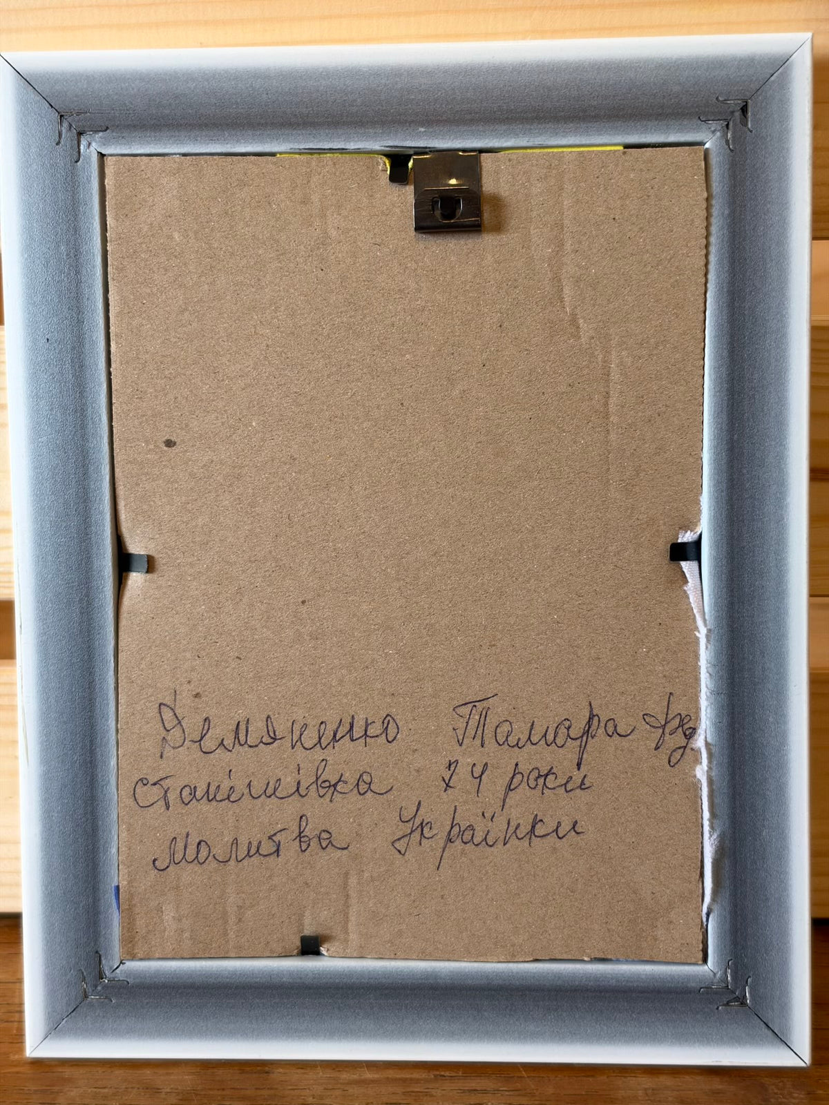 Reverse of the framed picture showing a brown backing board, retaining clips and a hanging fitting. Handwritten Ukrainian text identifies Tamara Demianenko, aged 74, Stanishivka, and the title “A Ukrainian Woman’s Prayer”.