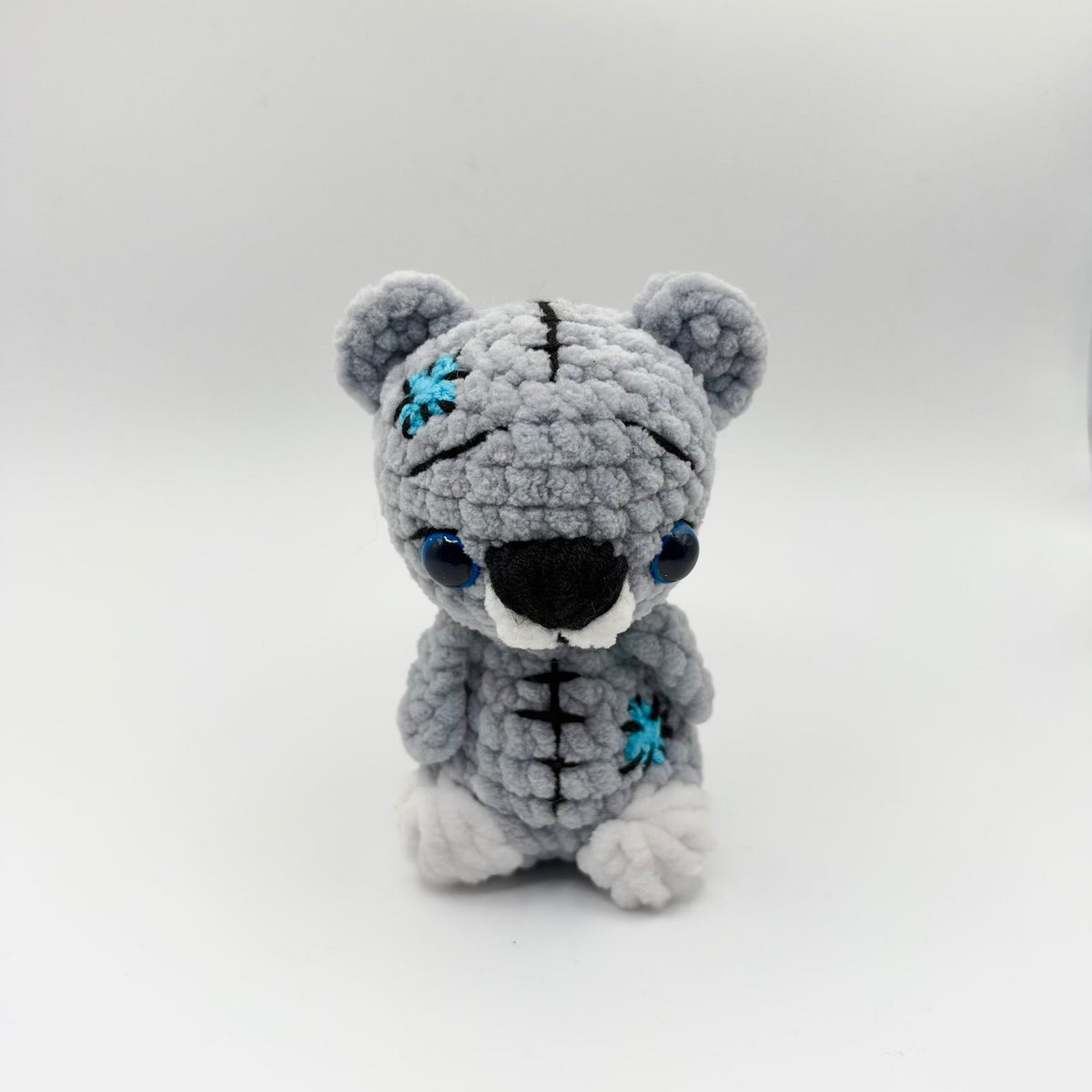 Small grey crochet teddy bear with large blue-rimmed eyes, a black nose, white muzzle and white paws. Black decorative stitching runs down its forehead and tummy, with turquoise stitched patches on its head and body.