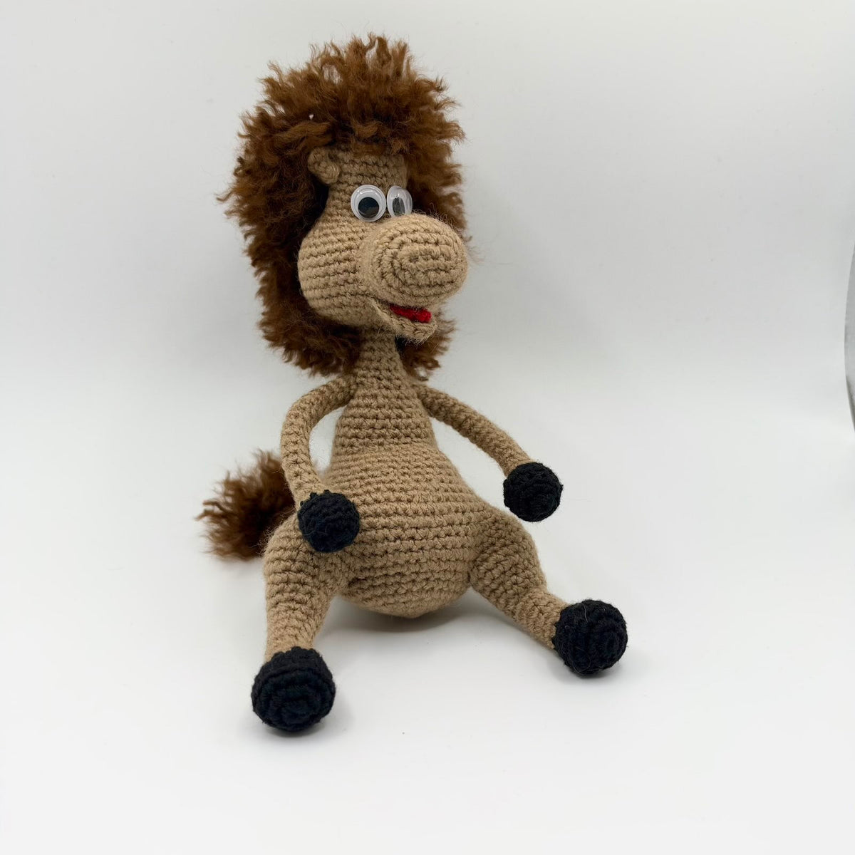 Beige crochet horse plushie sitting with its legs spread out, black hooves, large googly eyes and an open smiling mouth with a red tongue. It has a shaggy dark brown mane, matching fluffy tail and a large rounded muzzle.