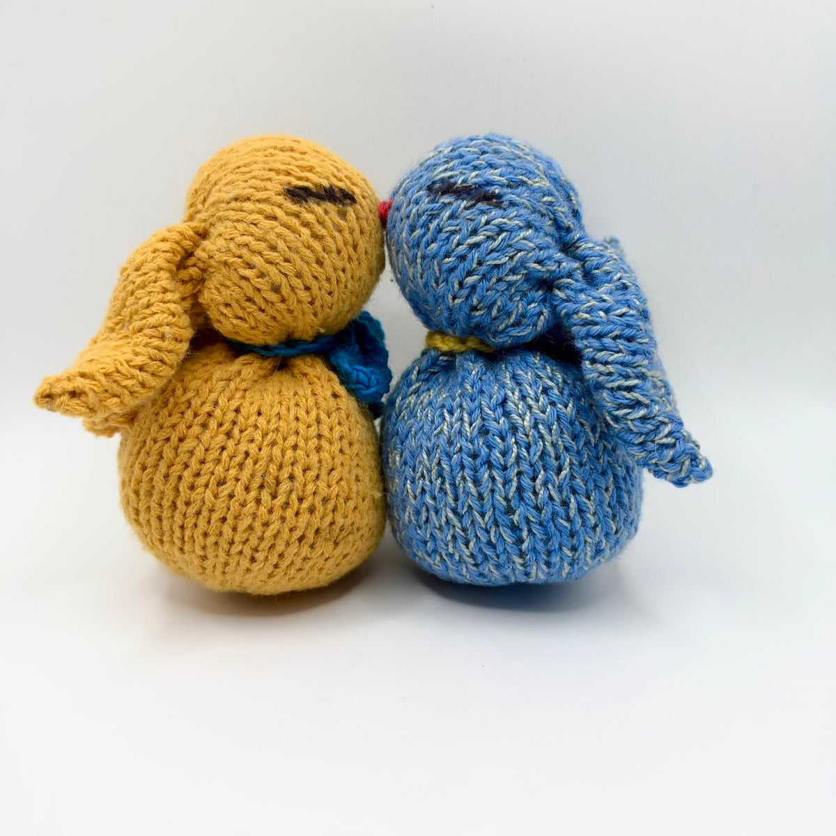 Two knitted bunny plushies nestled nose to nose, with long floppy ears, round bodies and closed embroidered eyes. The golden-yellow bunny wears a blue bow, and the flecked blue bunny wears a yellow neck tie.