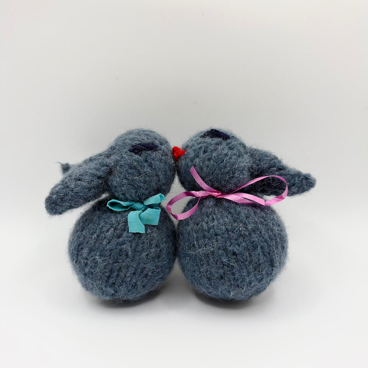 Two fuzzy grey knitted bunny plushies nestled nose to nose, with floppy ears, round bodies, closed embroidered eyes and red noses. One wears a turquoise fabric bow and the other a pink ribbon bow.