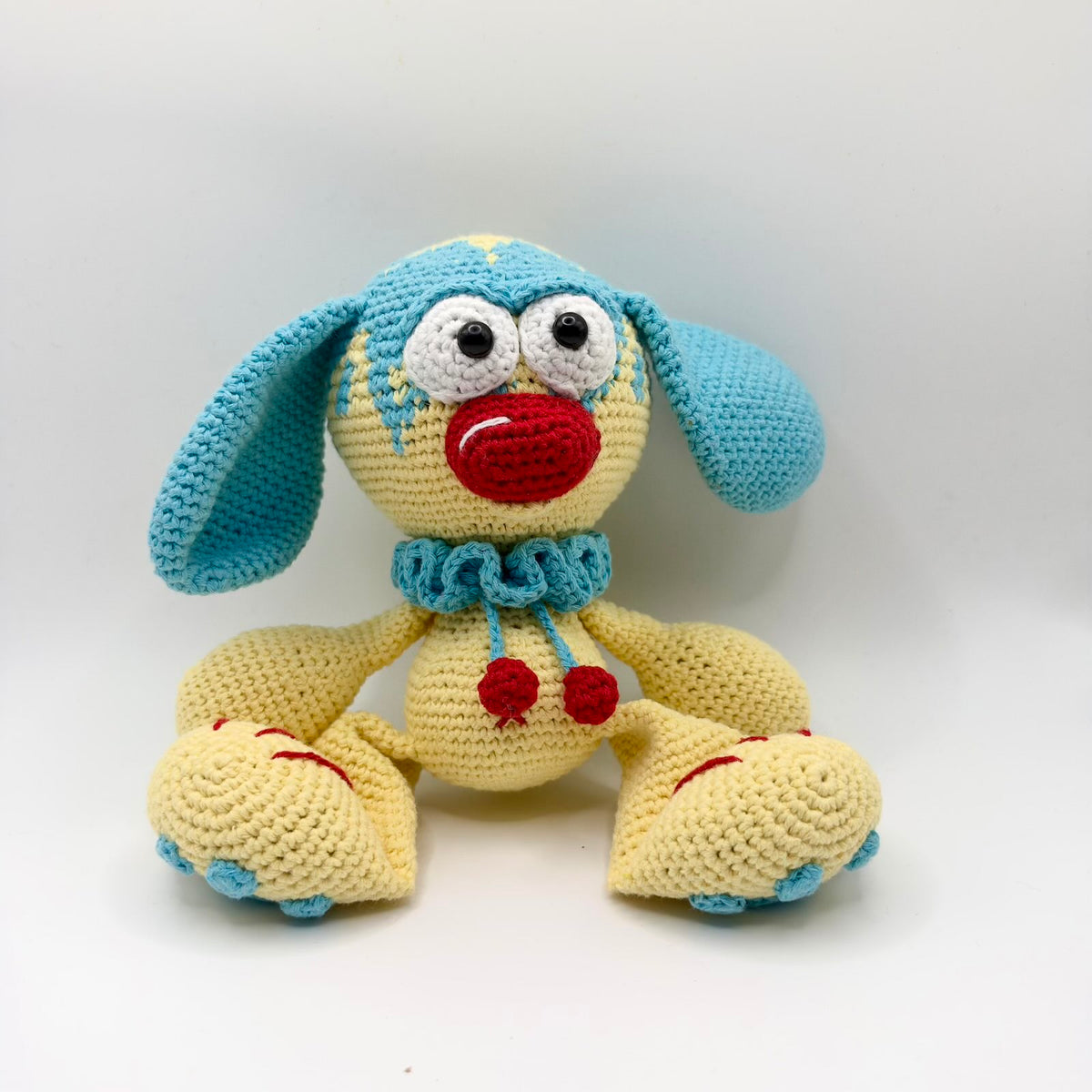 Pale yellow crochet character with long turquoise-blue floppy ears, blue detailing across its round head, large white eyes with black pupils and a bright red nose. It wears a blue ruffled collar with two dangling red bobbles and has oversized yellow feet with blue toe details and red stitching.