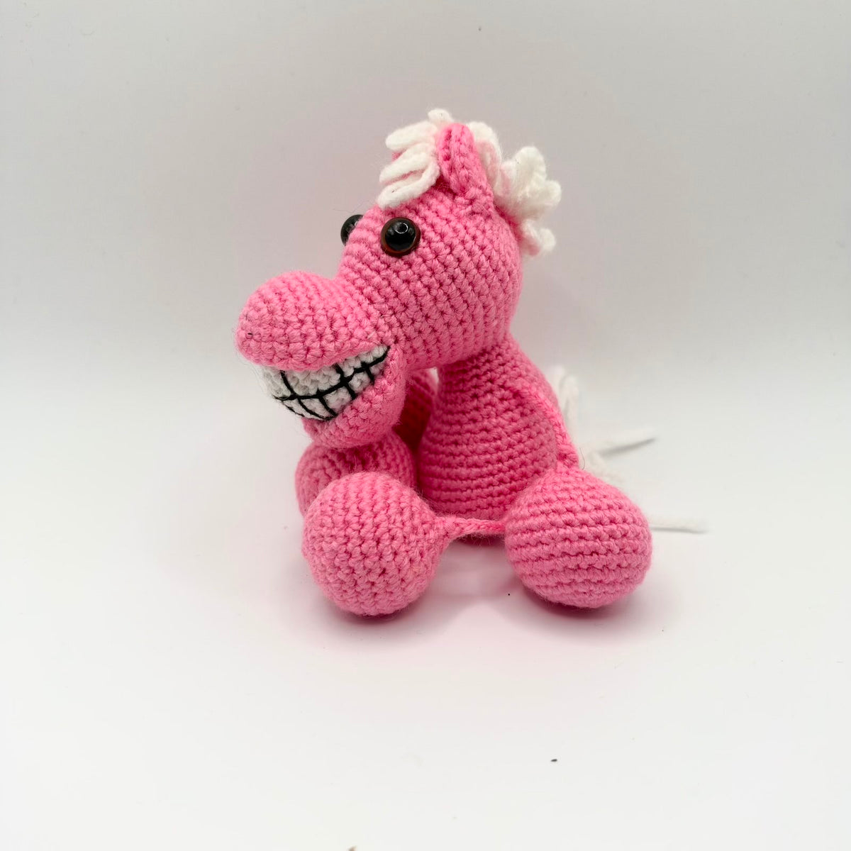 Bright pink crochet horse sitting at an angle against a white background, with a curly white yarn mane and white tail. It has upright ears, shiny dark eyes, a long rounded muzzle and a wide white grin divided into teeth with black stitching. Its legs end in large rounded pink hooves.