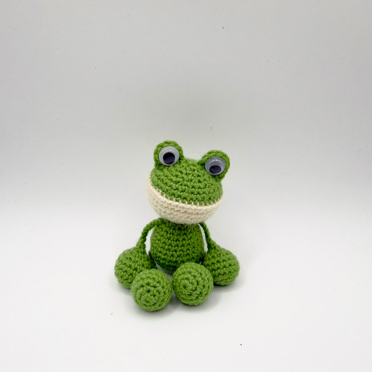 Bright green crochet frog sitting against a plain light background, with raised googly eyes and a cream-coloured lower face. Its small rounded body has thin green yarn limbs attached to large round crochet hands and feet, with visible spiral stitching on the feet.