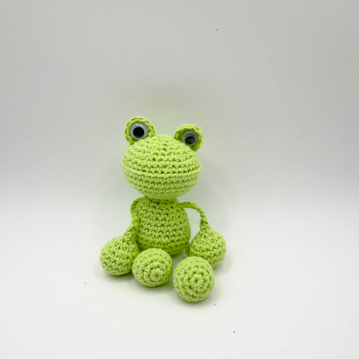 Lime-green crochet frog seated against a plain light background. It has a broad rounded head, raised googly eyes, a curved stitched mouth line and a small rounded body. Thin green yarn limbs connect to chunky round hands and feet, with spiral crochet stitches visible on the feet.