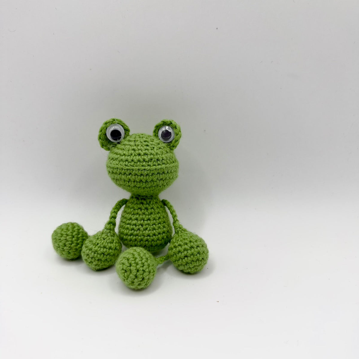 Leaf-green crochet frog with a large rounded head, raised googly eyes and a subtle curved mouth line. It has a small rounded body and thin yarn arms and legs ending in chunky rounded hands and feet. Seated with its limbs spread loosely against a plain light background.