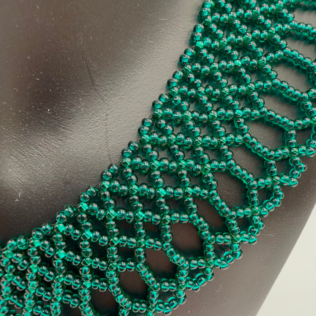 Close-up of translucent emerald-green beads forming the necklaceβs lace-like pattern against a black display bust. Rows of linked geometric openings lead into long rounded loops and small scallops along the lower edge.