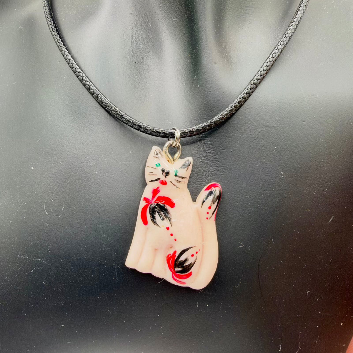 Glossy pale blush cat-shaped pendant with green eyes, black whiskers and a raised curved tail. Red-and-black floral motifs and small red dots decorate its body and tail. The pendant hangs from metal rings on a black braided cord, displayed on a black jewellery bust.