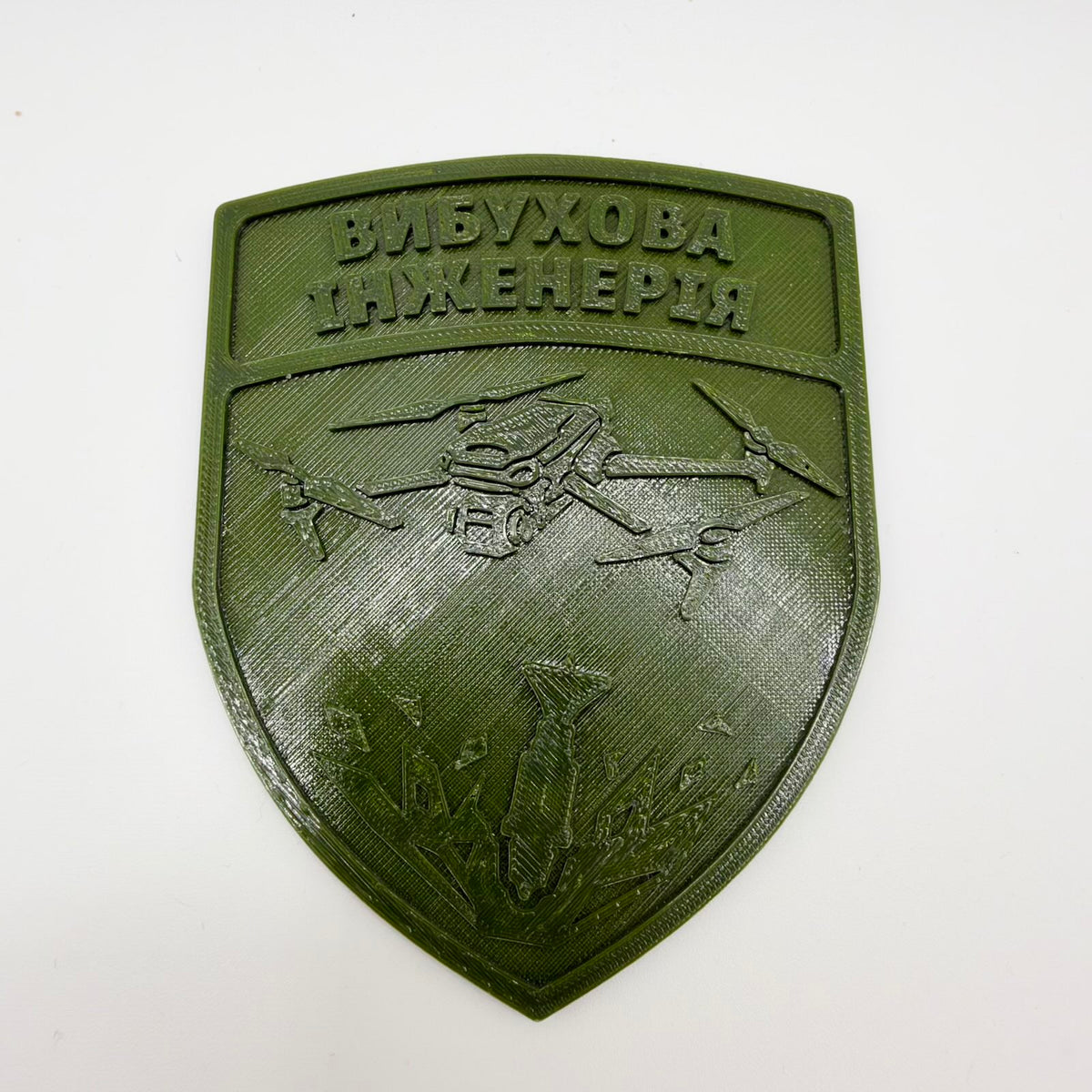 Olive-green 3D-printed shield with raised Ukrainian text “ВИБУХОВА ІНЖЕНЕРІЯ,” meaning “Explosive Engineering.” Below the lettering, a quadcopter appears above a falling bomb and stylised explosion. The shield has a raised border and visible diagonal print lines, photographed on white.