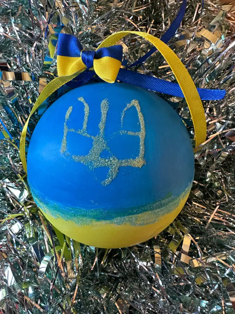 A hand painted Christmas bauble with a blue background and yellow and gold details, featuring a stylized face and a bow with blue and yellow ribbons.