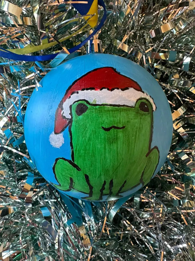 Hand painted Christmas bauble with a green frog wearing a Santa hat on a blue background, placed on a Christmas tree with silver glitter ornaments.