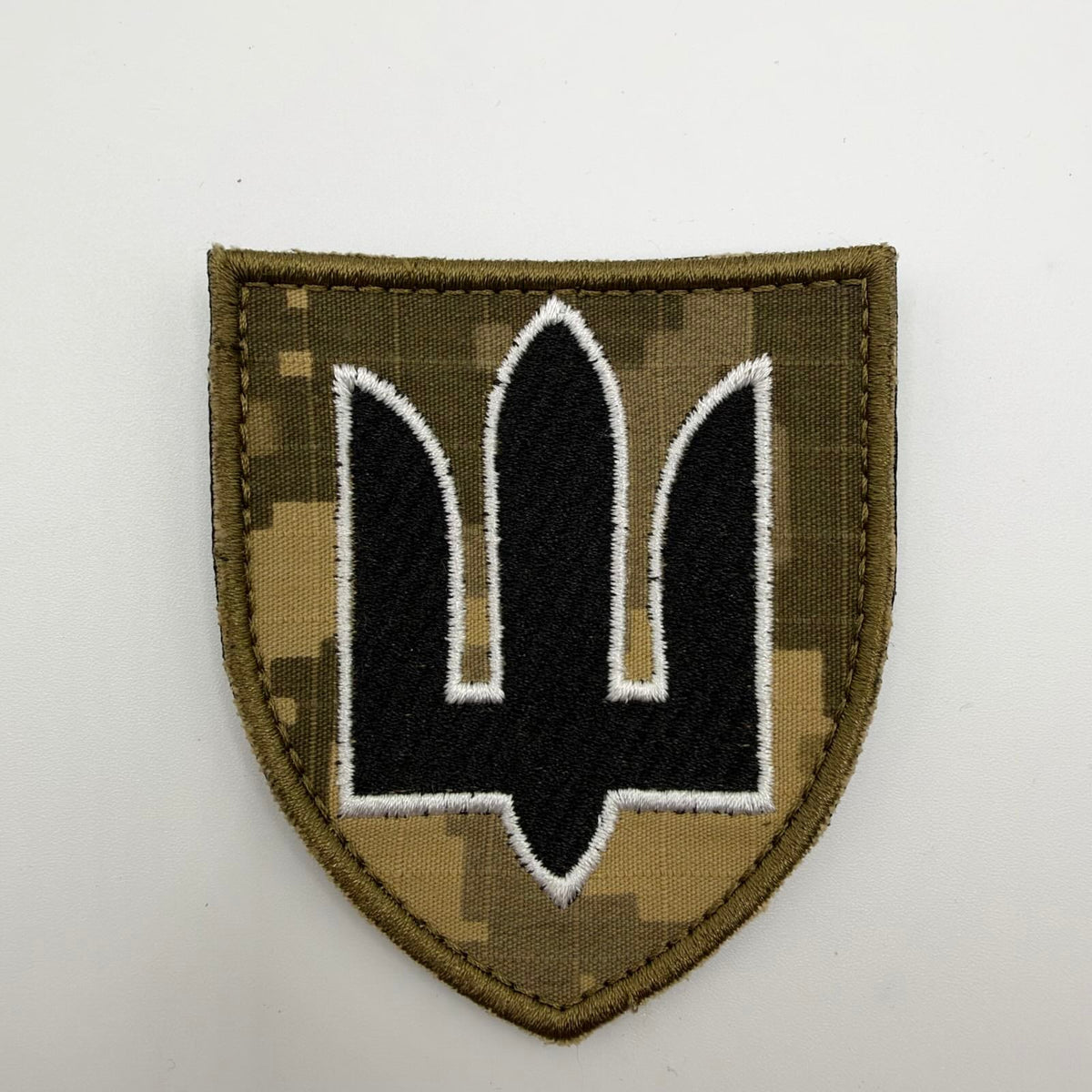 Shield-shaped Ukrainian MM-14 camouflage patch featuring a black embroidered stylised trident outlined in white, with an olive stitched border.