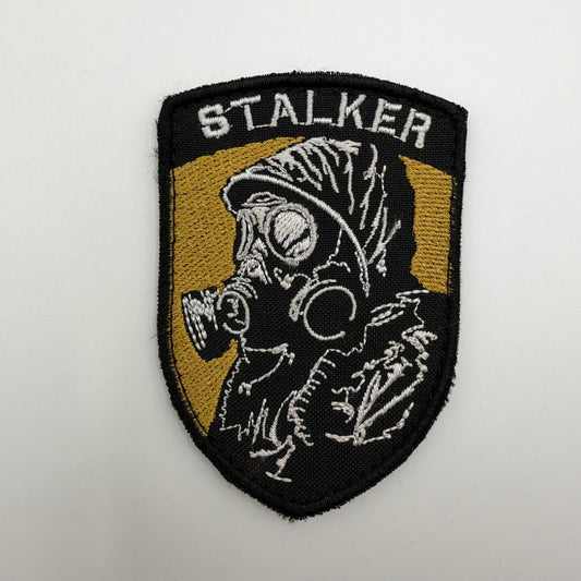 Shield-shaped STALKER embroidered patch featuring a hooded figure in a gas mask, stitched in white and grey against a gold and black background.