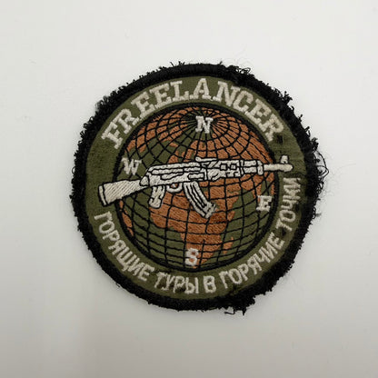 Worn circular Russian FREELANCER trophy patch with a rifle over an orange globe, Russian lettering and heavily frayed black edges.