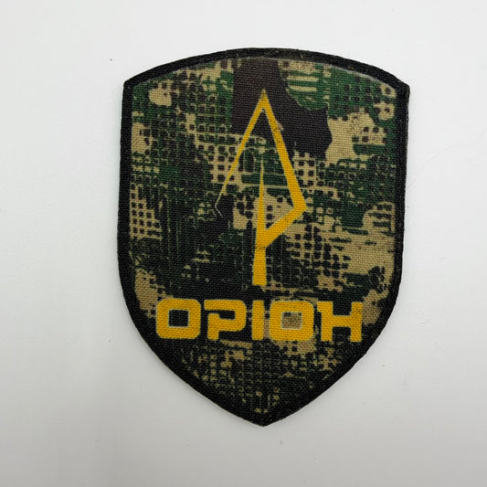 Shield-shaped Orion unit patch with a yellow spear-like emblem and Ukrainian “ОРІОН” lettering on green, tan, and black camouflage fabric.