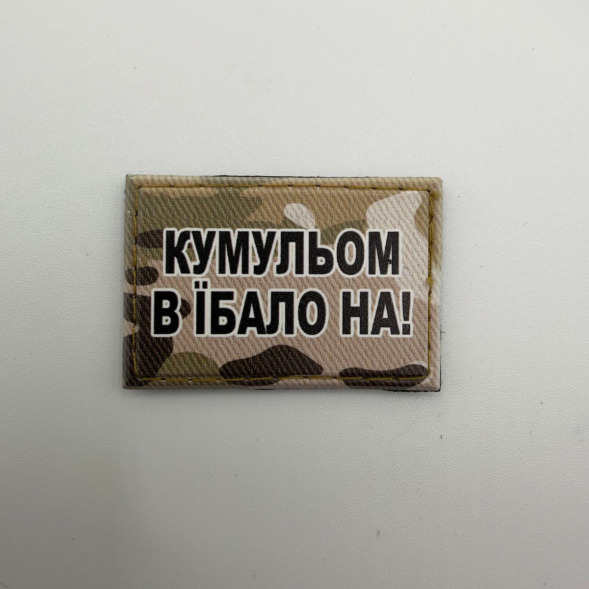Kristina’s rectangular camouflage morale patch with bold black, white-outlined Ukrainian lettering reading “КУМУЛЬОМ В ЇБАЛО НА!”