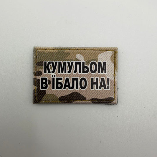 Kristina’s rectangular camouflage morale patch with bold black, white-outlined Ukrainian lettering reading “КУМУЛЬОМ В ЇБАЛО НА!”