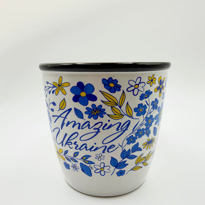White mug with a black rim, decorated with blue and yellow flowers and leaves surrounding blue “Amazing Ukraine” lettering.