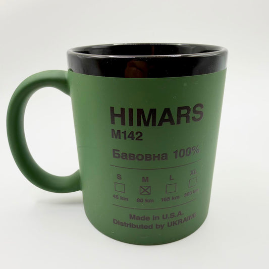 Military-green HIMARS M142 mug with a black interior and rim, featuring a clothing-label-style design and “Distributed by UKRAINE” wording.