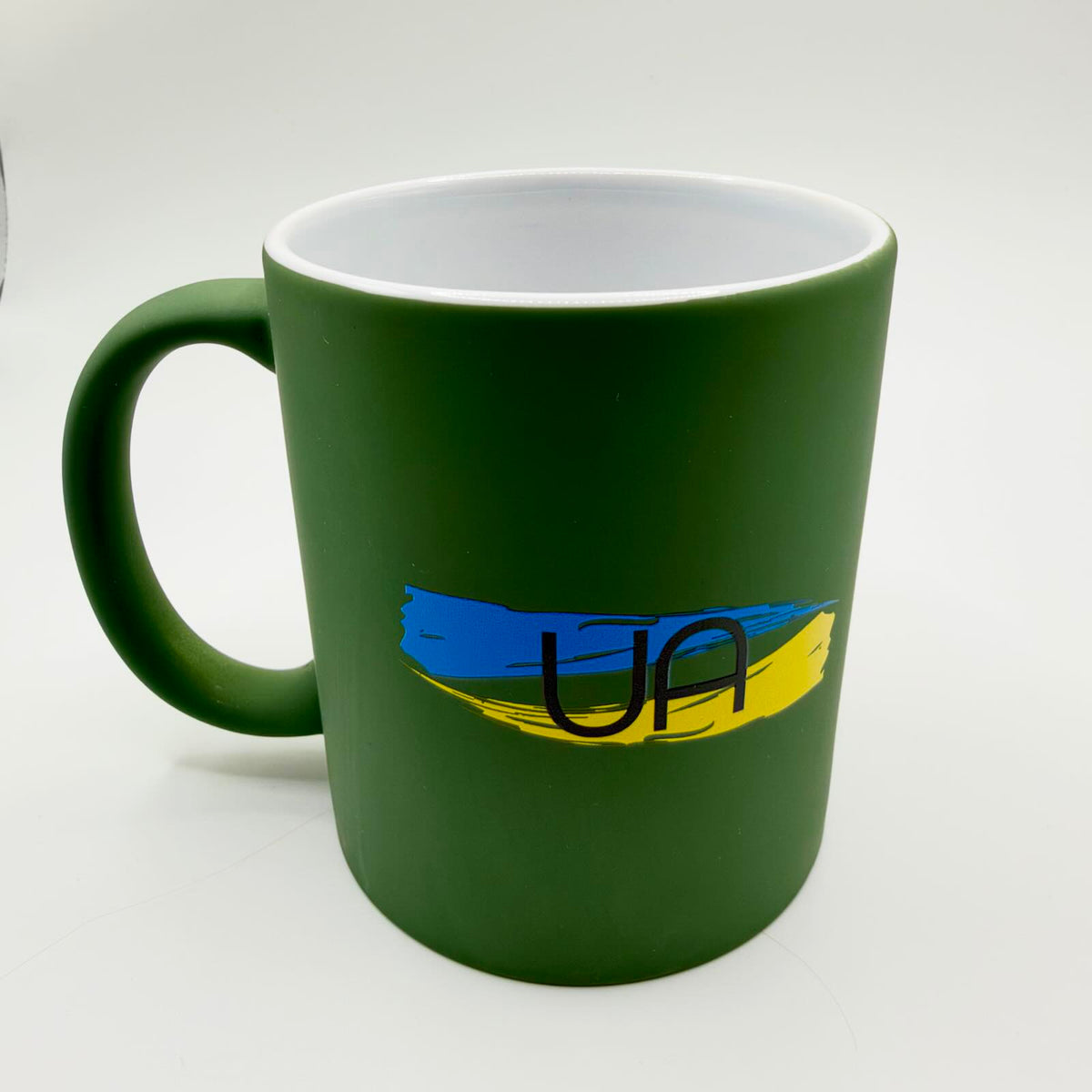 Military-green mug with a white interior and matching green handle, featuring black UA lettering over blue and yellow brushstrokes.