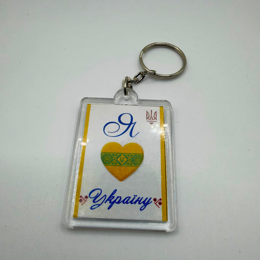 Clear rectangular keyring with blue Ukrainian “I love Ukraine” lettering, a patterned yellow heart, trident details and a metal split ring.