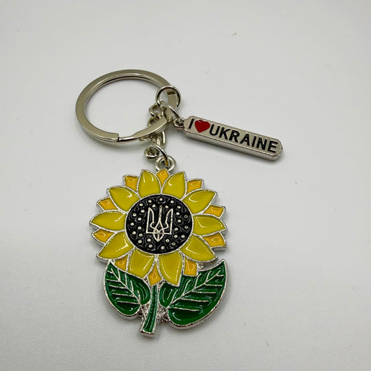 Silver-coloured keyring with a yellow sunflower charm, green leaves and a Ukrainian trident in its black centre, alongside a small “I love Ukraine” tag with a red heart.