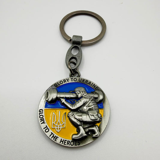 Round silver-tone keyring featuring a soldier with a Javelin launcher, Ukrainian flag colours, trident and “Glory to Ukraine, Glory to the Heroes” wording.
