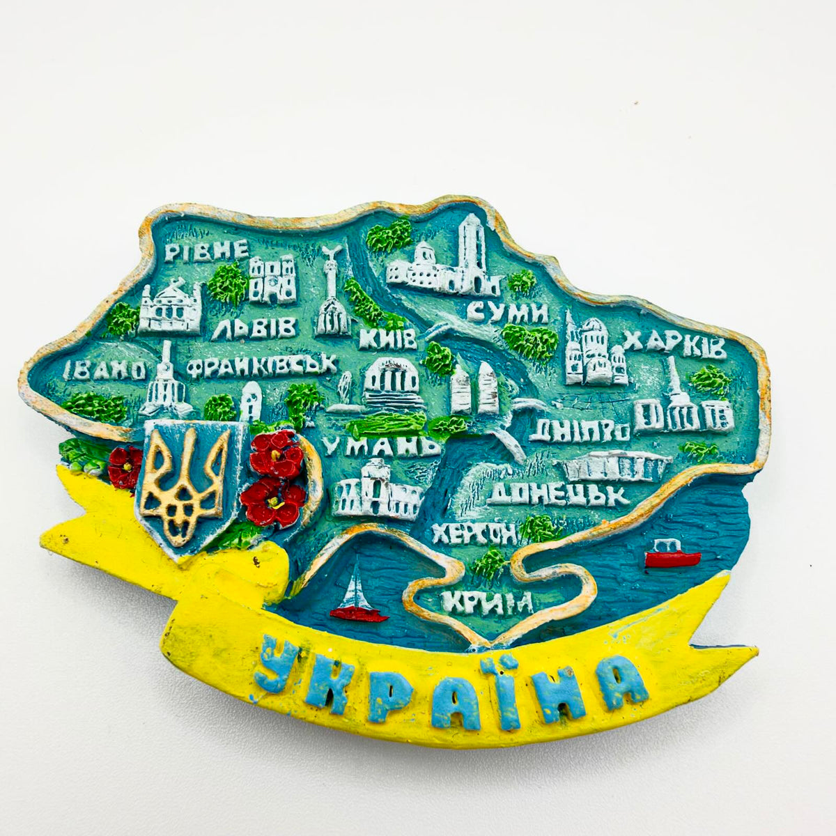 Relief-style Ukraine map fridge magnet including Crimea, with labelled cities, miniature landmarks, a Tryzub shield, red flowers and a yellow “Ukraine” ribbon.