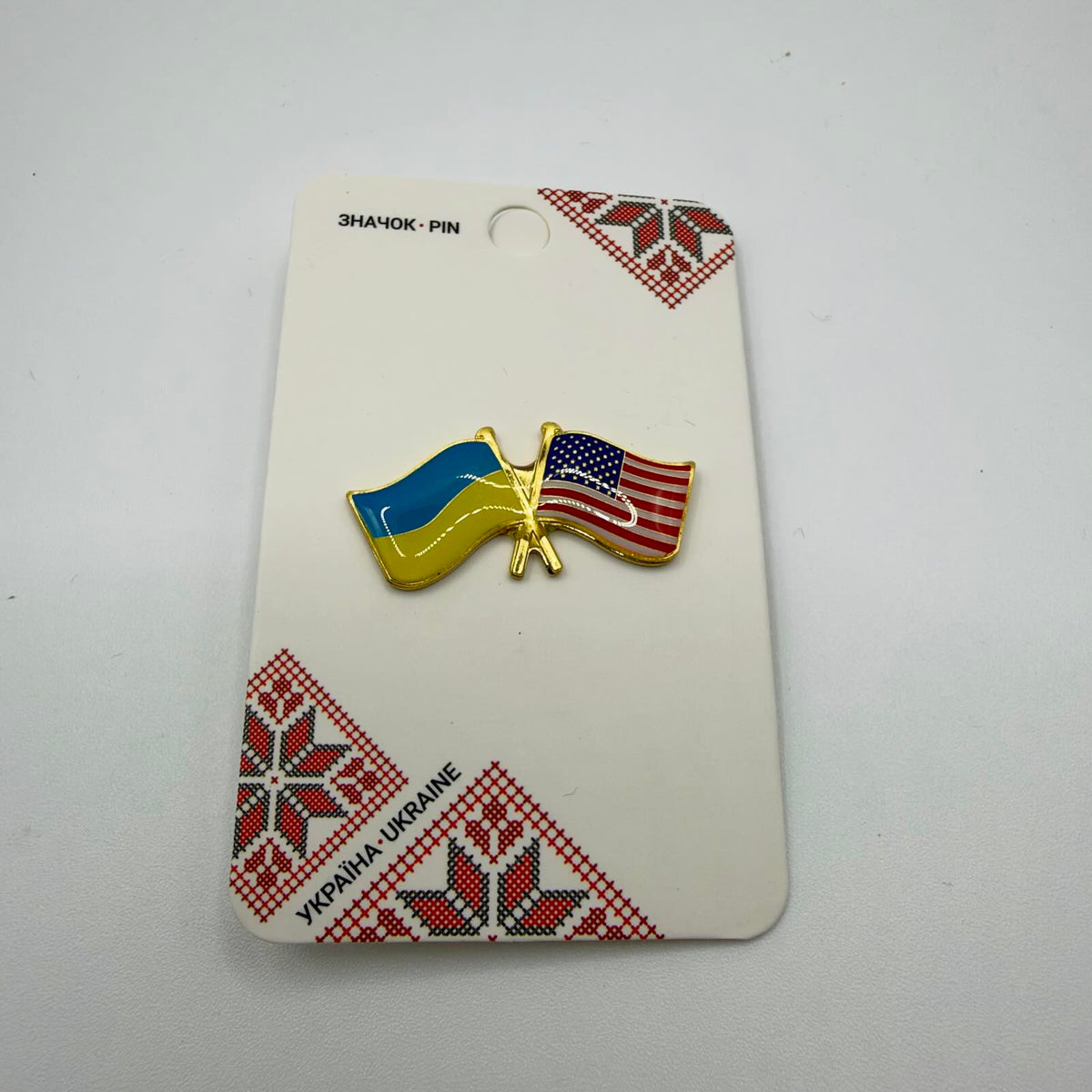 Gold-coloured crossed-flags pin featuring the Ukrainian and United States flags, mounted on a card with red and black Ukrainian embroidery-style patterns.
