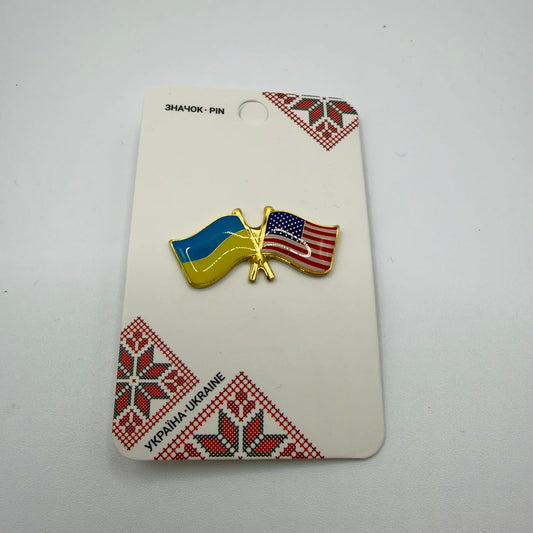 Gold-coloured crossed-flags pin featuring the Ukrainian and United States flags, mounted on a card with red and black Ukrainian embroidery-style patterns.