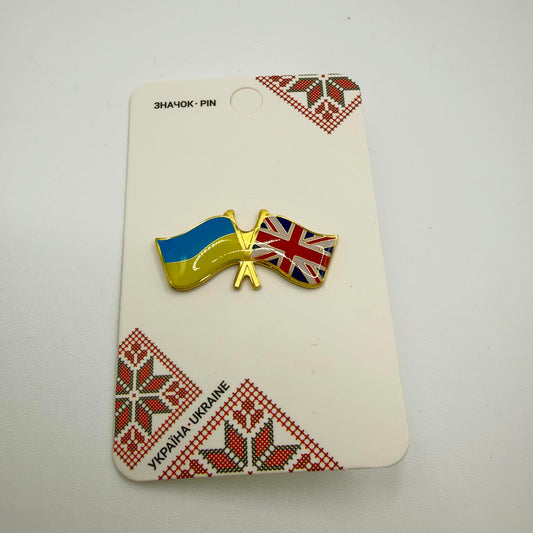 Gold-coloured crossed-flags pin featuring the Ukrainian flag and British Union Flag, mounted on a card with red and black Ukrainian embroidery-style patterns.