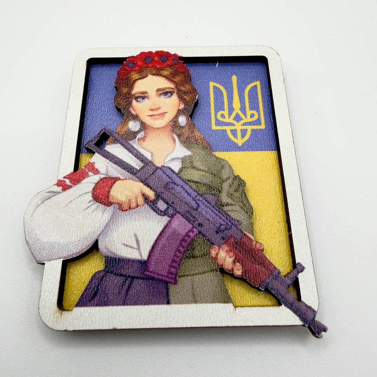 Layered fridge magnet depicting a woman with a red flower wreath, embroidered blouse and military-style jacket holding a rifle, against a Ukrainian flag and yellow Tryzub.