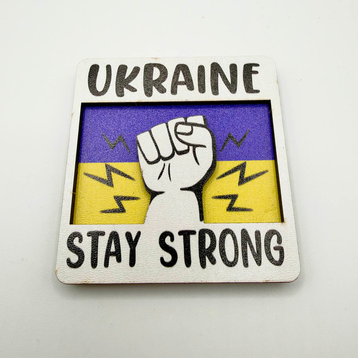 Layered fridge magnet with a raised clenched fist against a blue and yellow background, framed by the words “Ukraine — Stay Strong”.