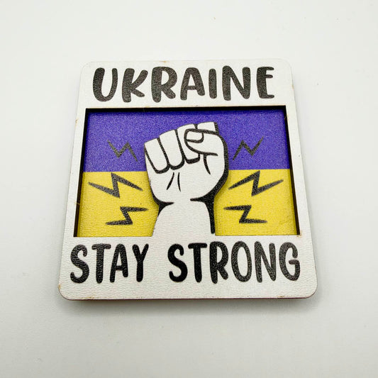 Layered fridge magnet with a raised clenched fist against a blue and yellow background, framed by the words “Ukraine — Stay Strong”.