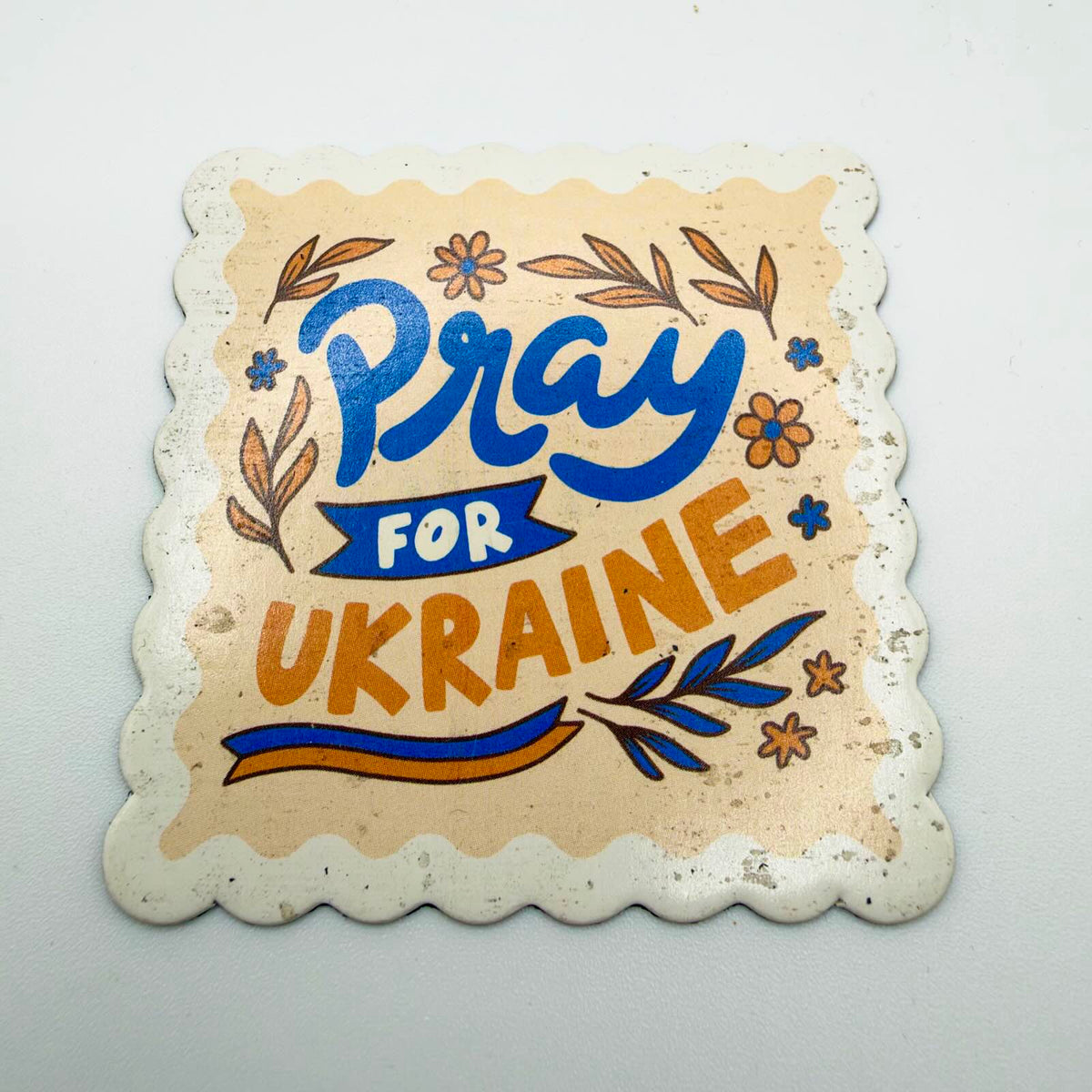 Scalloped rectangular fridge magnet reading “Pray for Ukraine” in blue and golden-yellow lettering, surrounded by flowers, leaves and a ribbon on a cream background.
