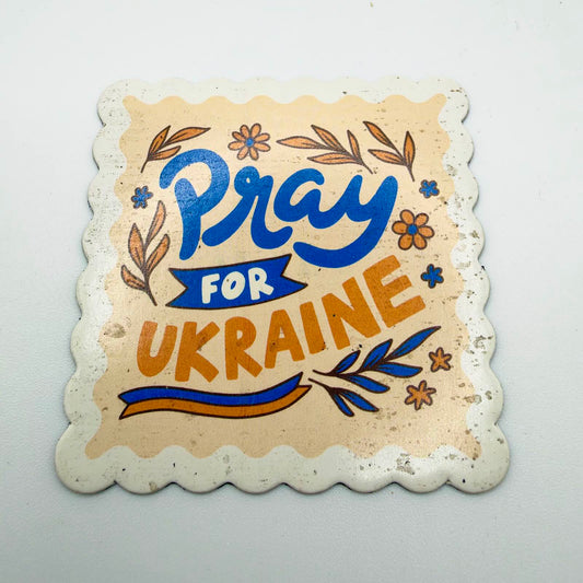 Scalloped rectangular fridge magnet reading “Pray for Ukraine” in blue and golden-yellow lettering, surrounded by flowers, leaves and a ribbon on a cream background.