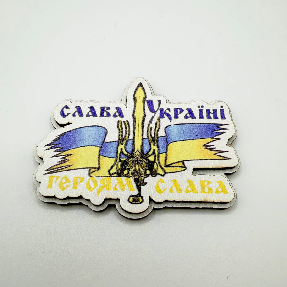 Shaped fridge magnet with a Ukrainian blue and yellow ribbon, central trident-and-sword motif, and Ukrainian lettering meaning “Glory to Ukraine — Glory to the Heroes”.