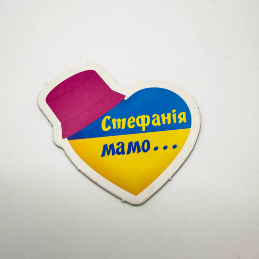 Heart-shaped fridge magnet in Ukrainian blue and yellow, topped with a pink bucket hat and Ukrainian text reading “Stefania, Mum…”