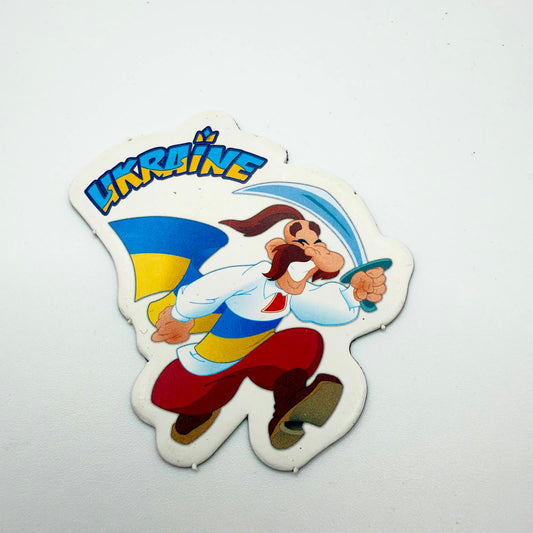 Shaped fridge magnet showing a smiling cartoon Cossack holding a curved sabre, with red trousers, a Ukrainian flag and blue and yellow “UKRAINE” lettering.