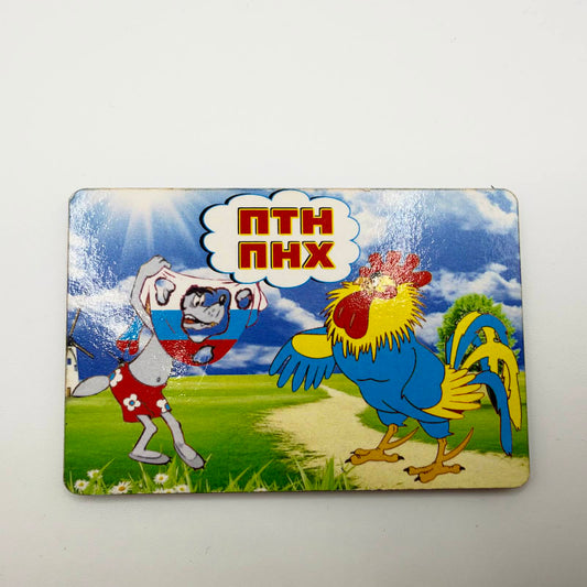 Cartoon fridge magnet showing a blue and yellow rooster facing a wolf tangled in Russian flag colours, beneath “ПТН ПНХ” lettering.
