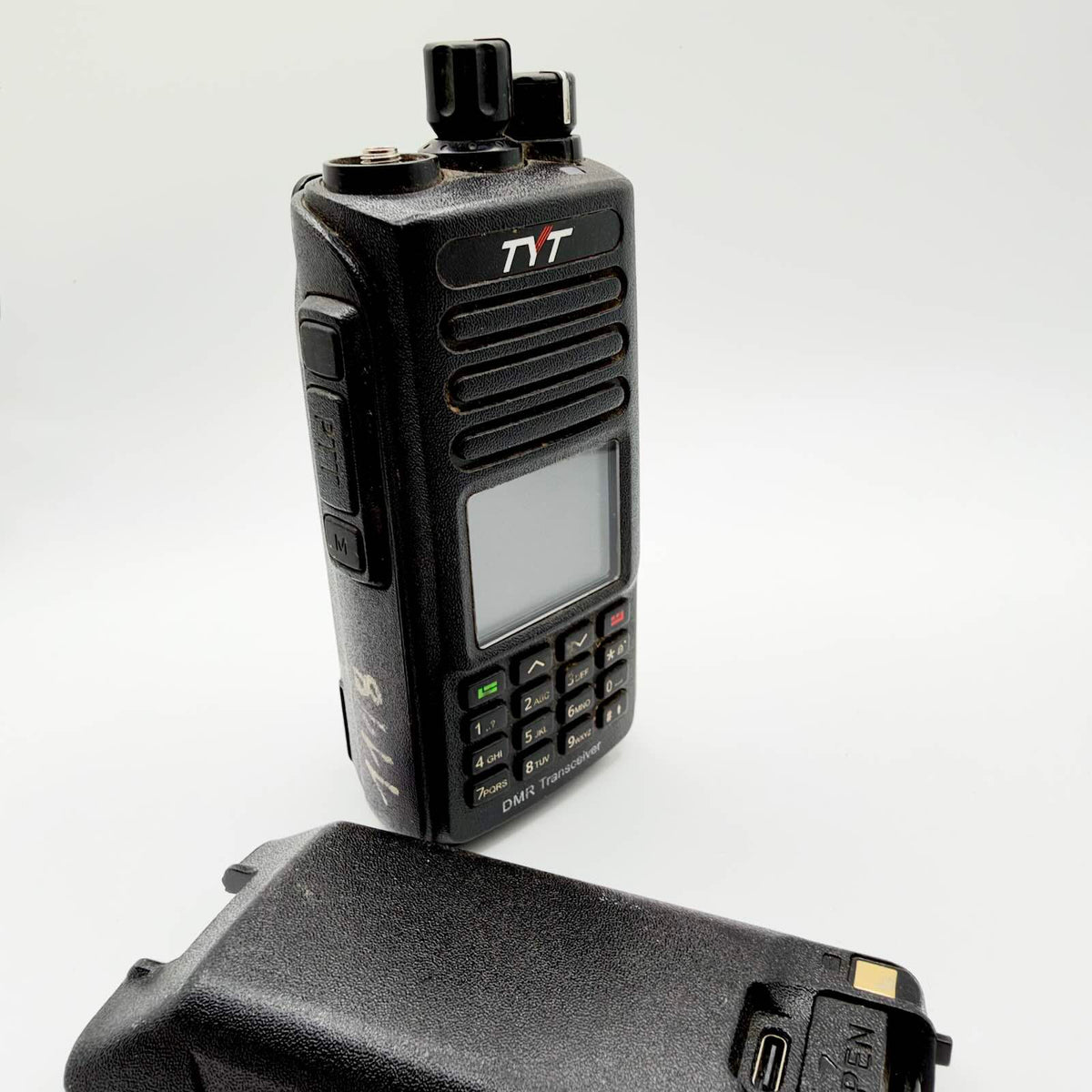 Black TYT handheld digital radio with keypad, missing antenna and detached battery on a white background.