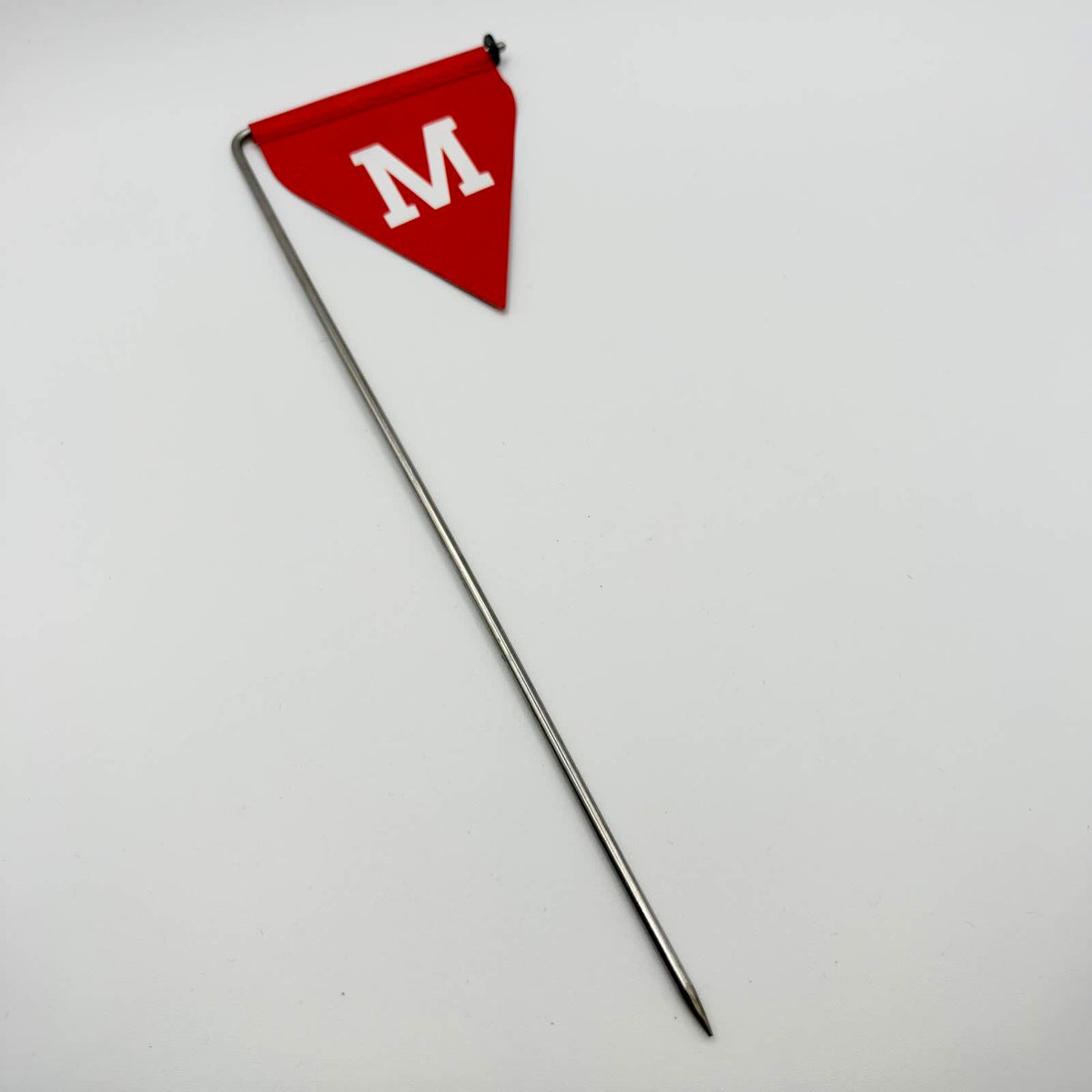Full-length view of a red triangular mine warning flag with a white “М”, mounted on a long, pointed metal stake against a white background.