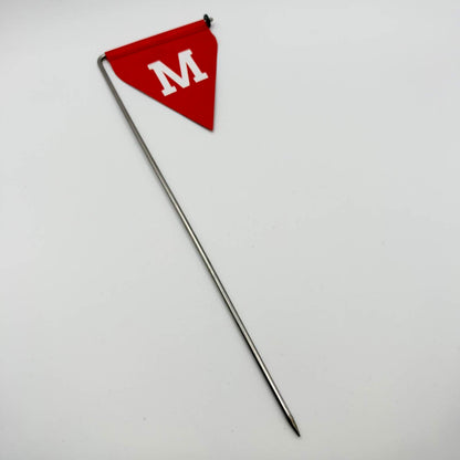 Full-length view of a red triangular mine warning flag with a white “М”, mounted on a long, pointed metal stake against a white background.