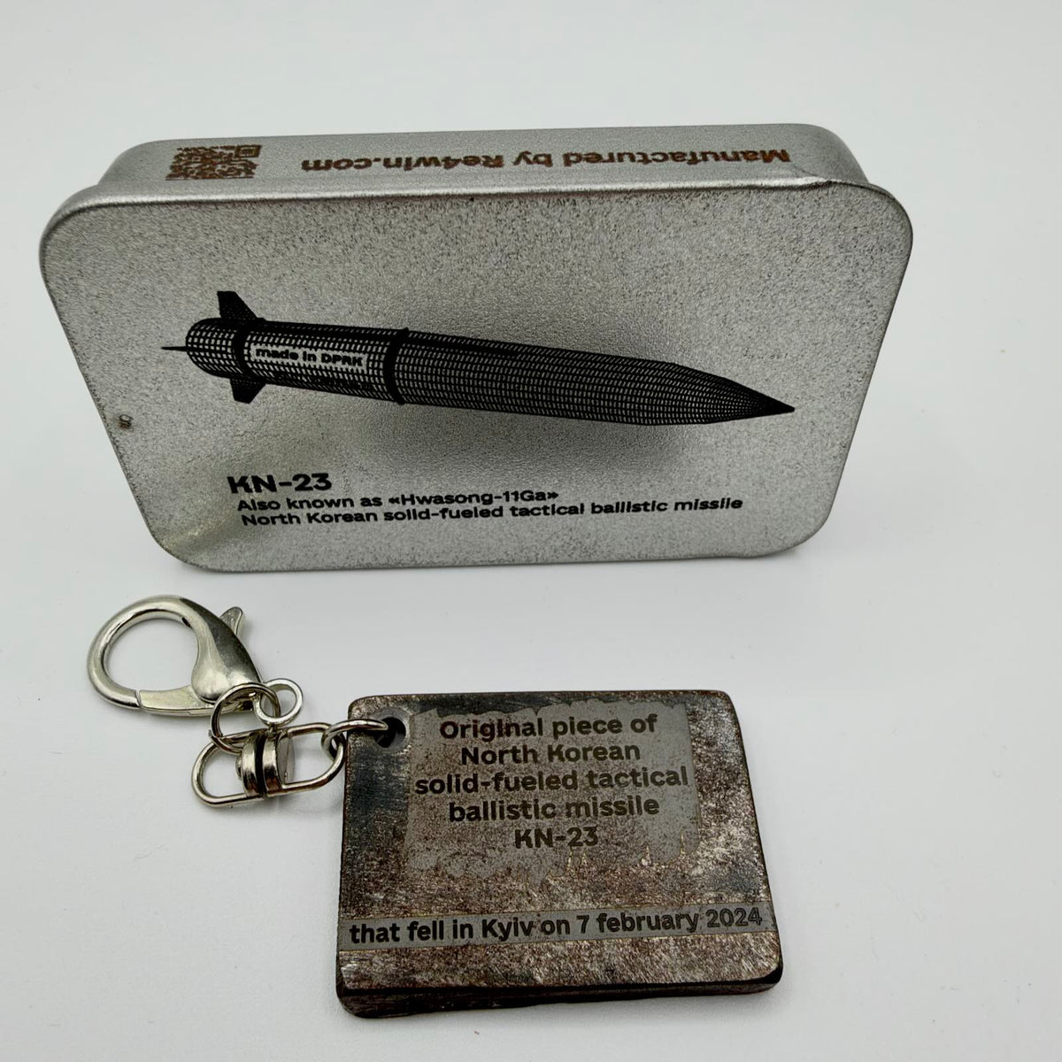 Rectangular KN-23 missile fragment keyring inscribed with Kyiv, 7 February 2024, beside a RE4WIN presentation tin showing the missile.